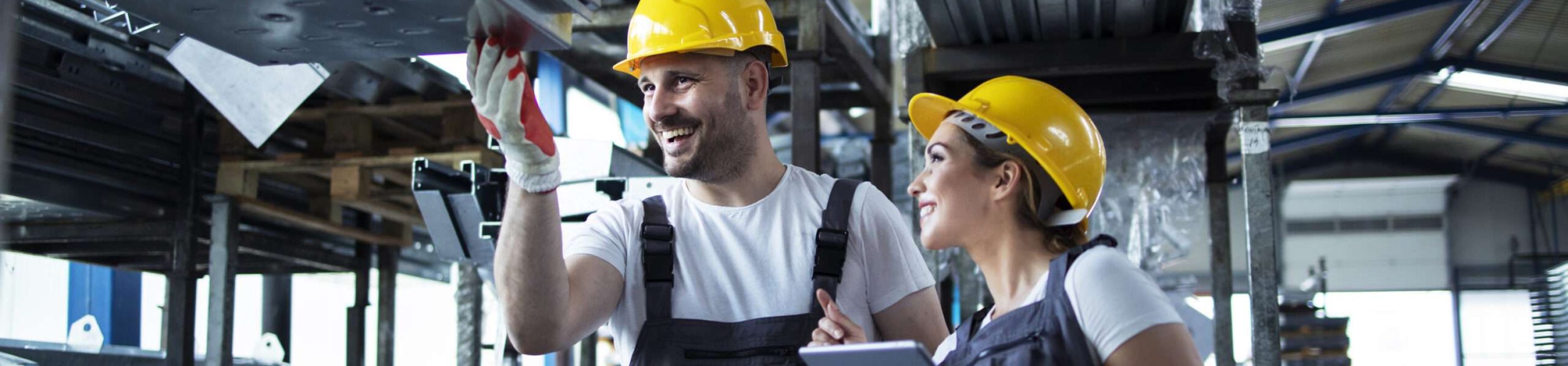 Factory workers checking inventory with tablet computer in industrial warehouse full of metal parts.