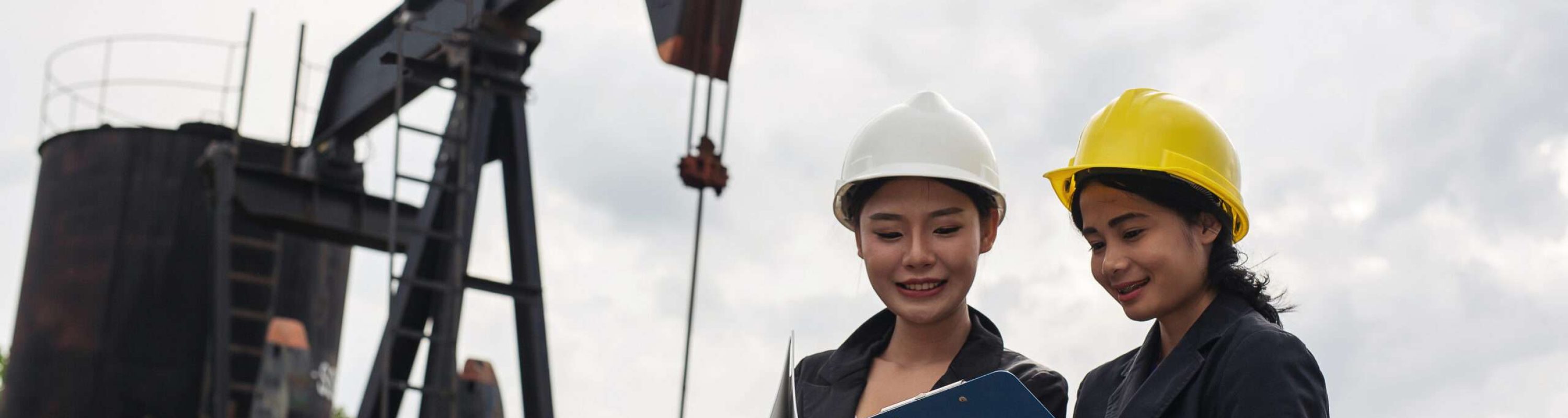 Two female engineers stand beside working oil pumps with a white sky background.